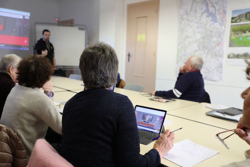 Autour d'une grande table, des personnes écoutent avec un ordinateur ou une tablette devant elles. Un animateur, debout, explique quelque chose devant un tableau blanc.